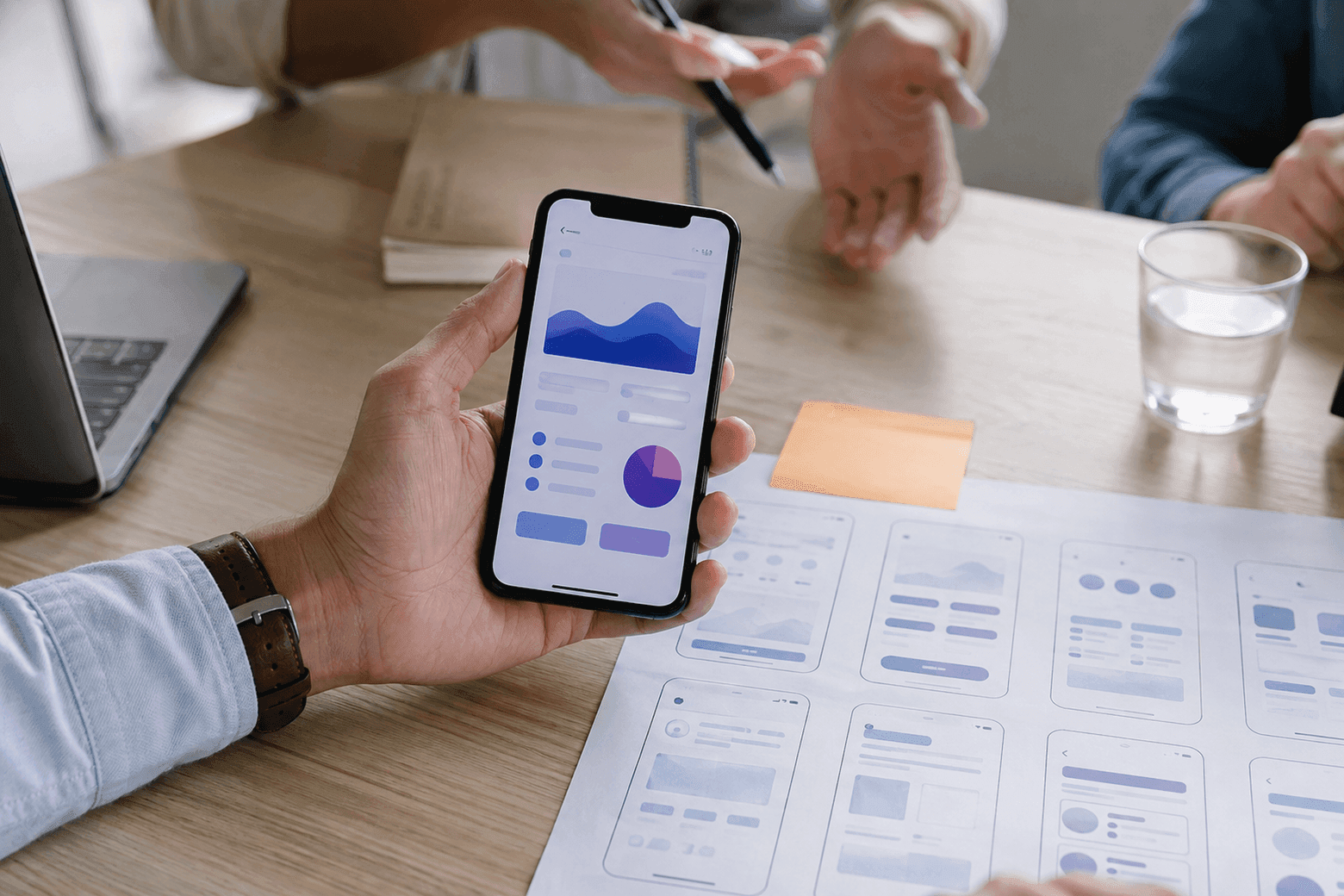 Une main tient un iPhone avec l'application ouverte au-dessus d'une table couverte de maquettes d'écrans imprimées
